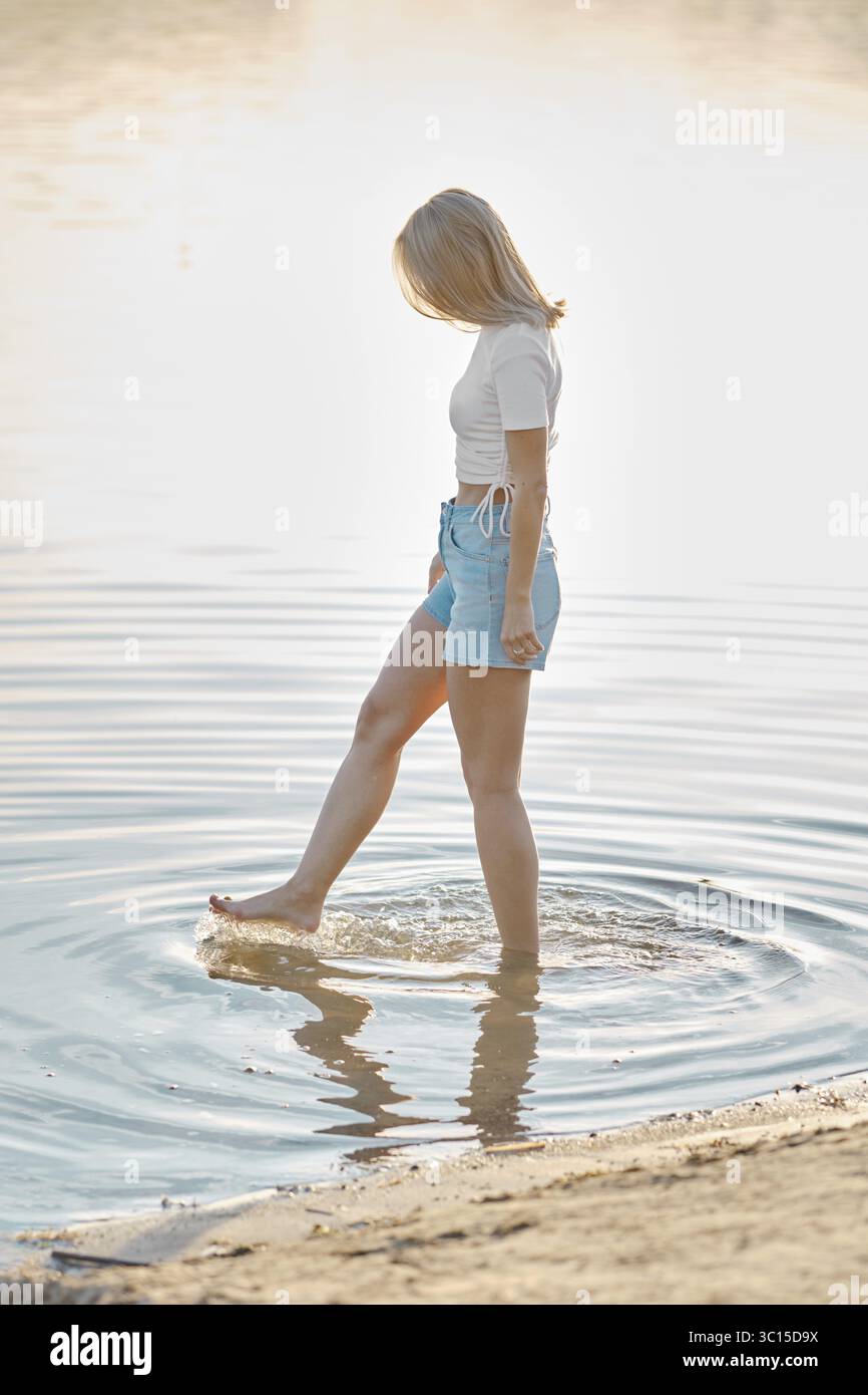 A woman stands in shallow water at a beach during golden hour ...