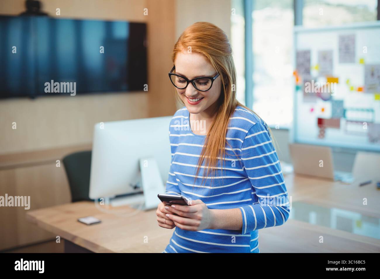 Red-haired woman standing by wooden office desk checking smartphone with monitor and whiteboard Stock Photo