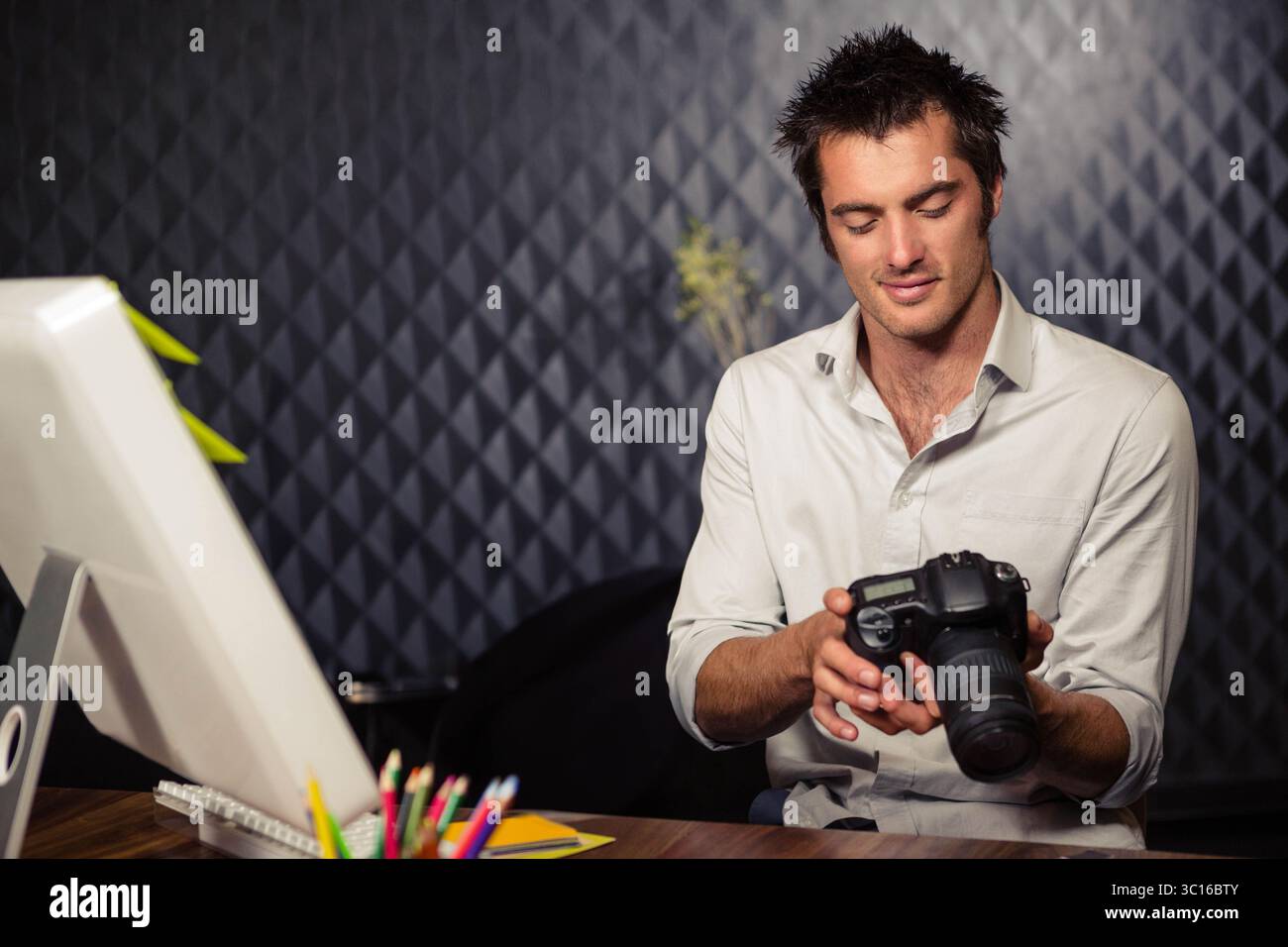 Man examining DSLR camera while sitting at wooden desk with computer monitor and colored pencils Stock Photo