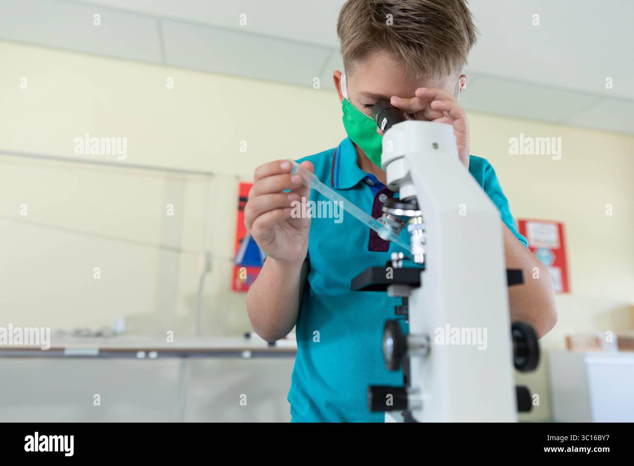 Boy wearing green mask peering into white microscope holding pipette ...