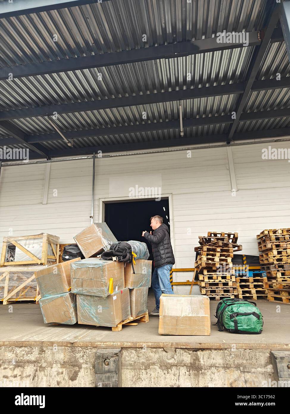 Individual unloading packages from truck at warehouse, surrounded by stacked pallets and storage area, showcasing logistics operations Stock Photo
