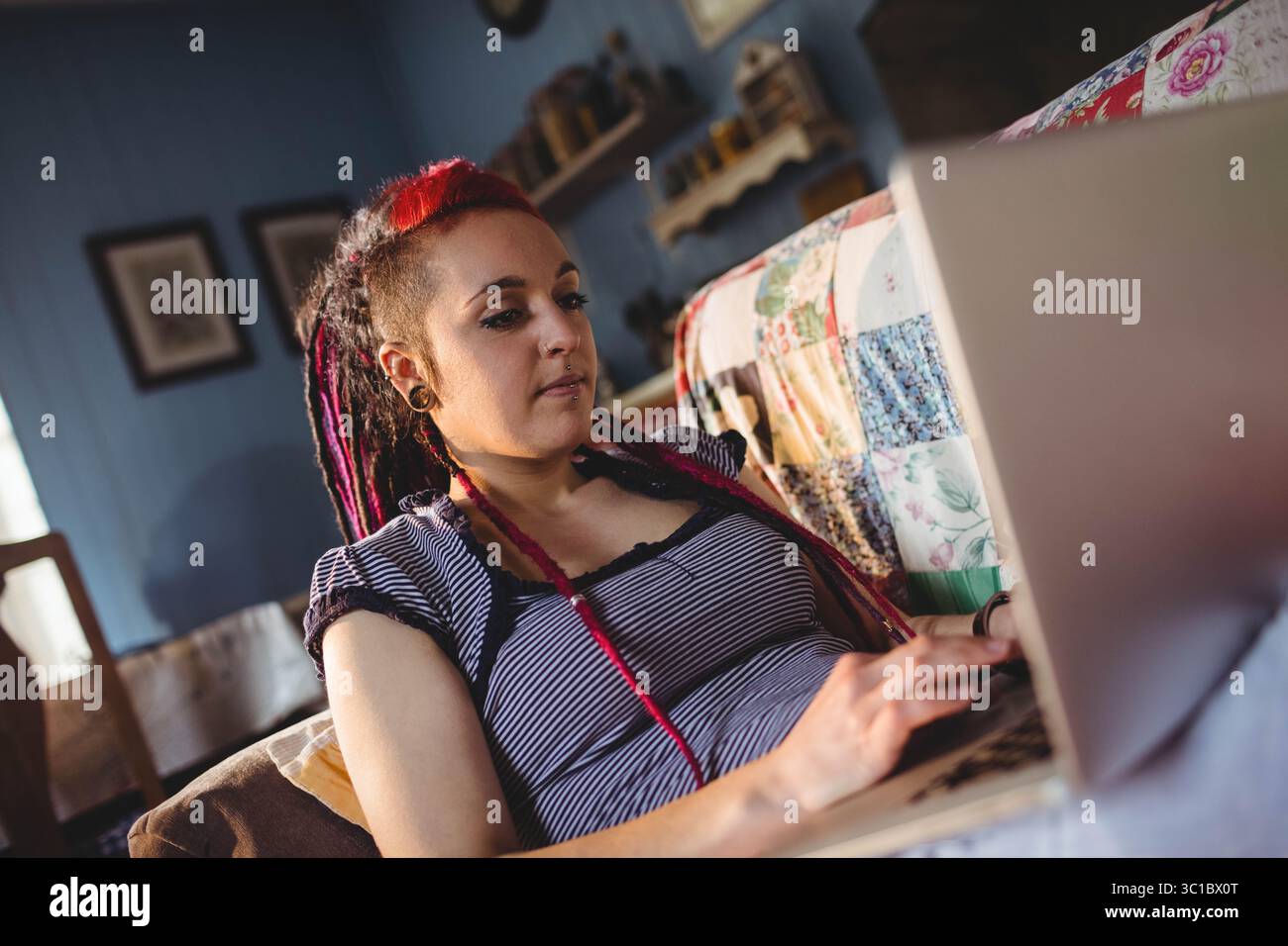 Woman reclining on patchwork quilt sofa in living room, using laptop with natural light Stock Photo