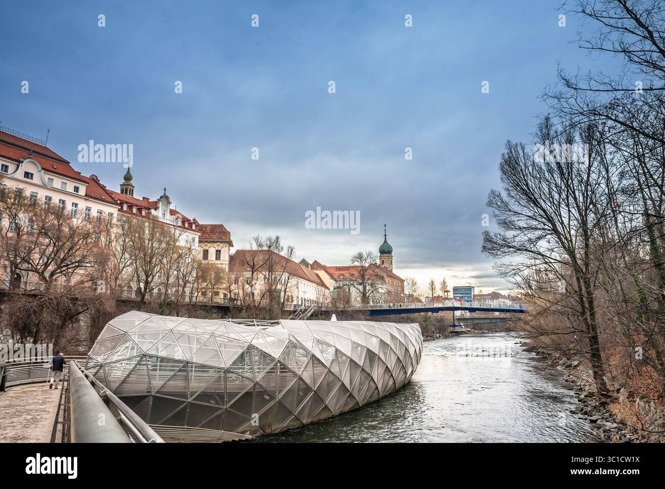 GRAZ, AUSTRIA - DECEMBER 17, 2024: The glass panelled Murinsel pavilion floats in the Mur river at Graz, Austria, as a solitary walker crosses the bri Stock Photo