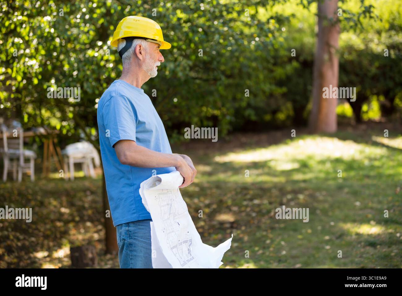 yellow hard hat resting beside blueprints on grassy lawn in filtered sunlight with white chairs Stock Photo
