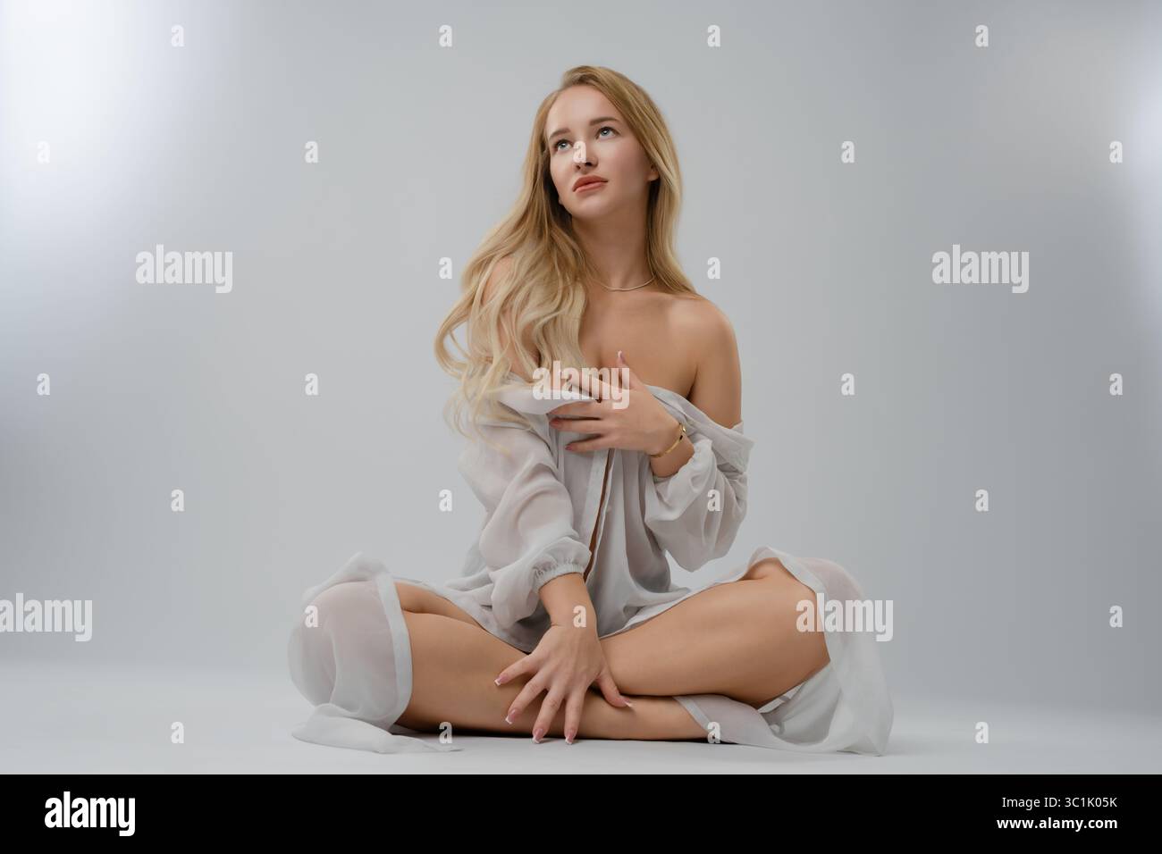Blonde woman in studio poses elegantly with soft expression and flowing attire Stock Photo