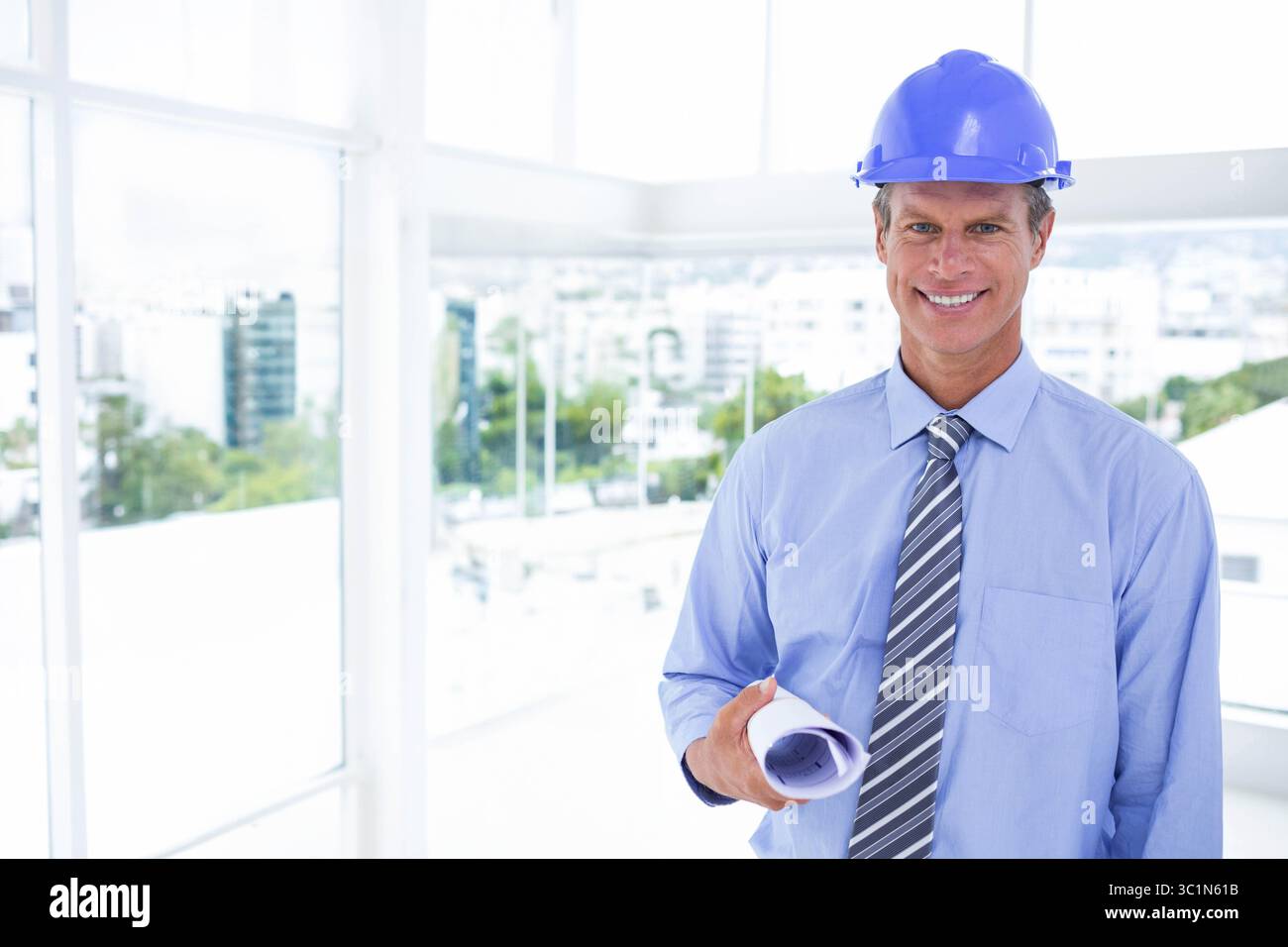Middle-aged man wearing blue hard hat holding rolled blueprints standing inside office, copy space Stock Photo