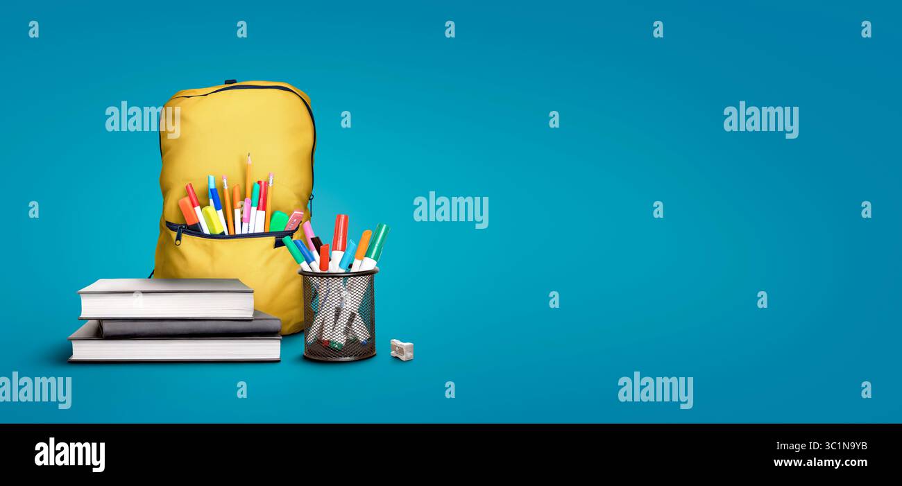 A back to school scene of a pile of three study books to learn from, yellow school bag and pens and pencils against a blue background with copy space. Stock Photo