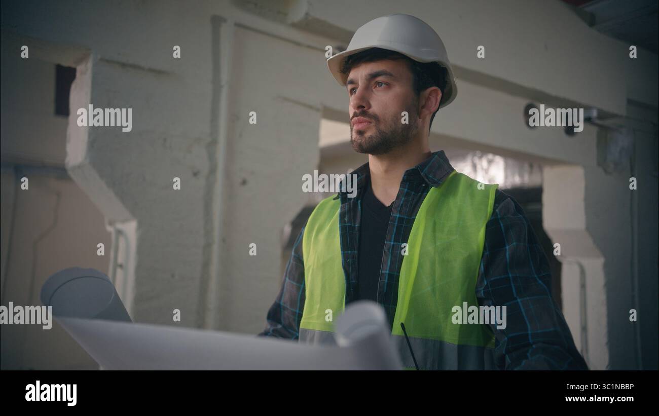 Caucasian man guy builder unfold blueprint in construction engineer serious architect looking at drawing draft paper manufactory male inspect Stock Photo