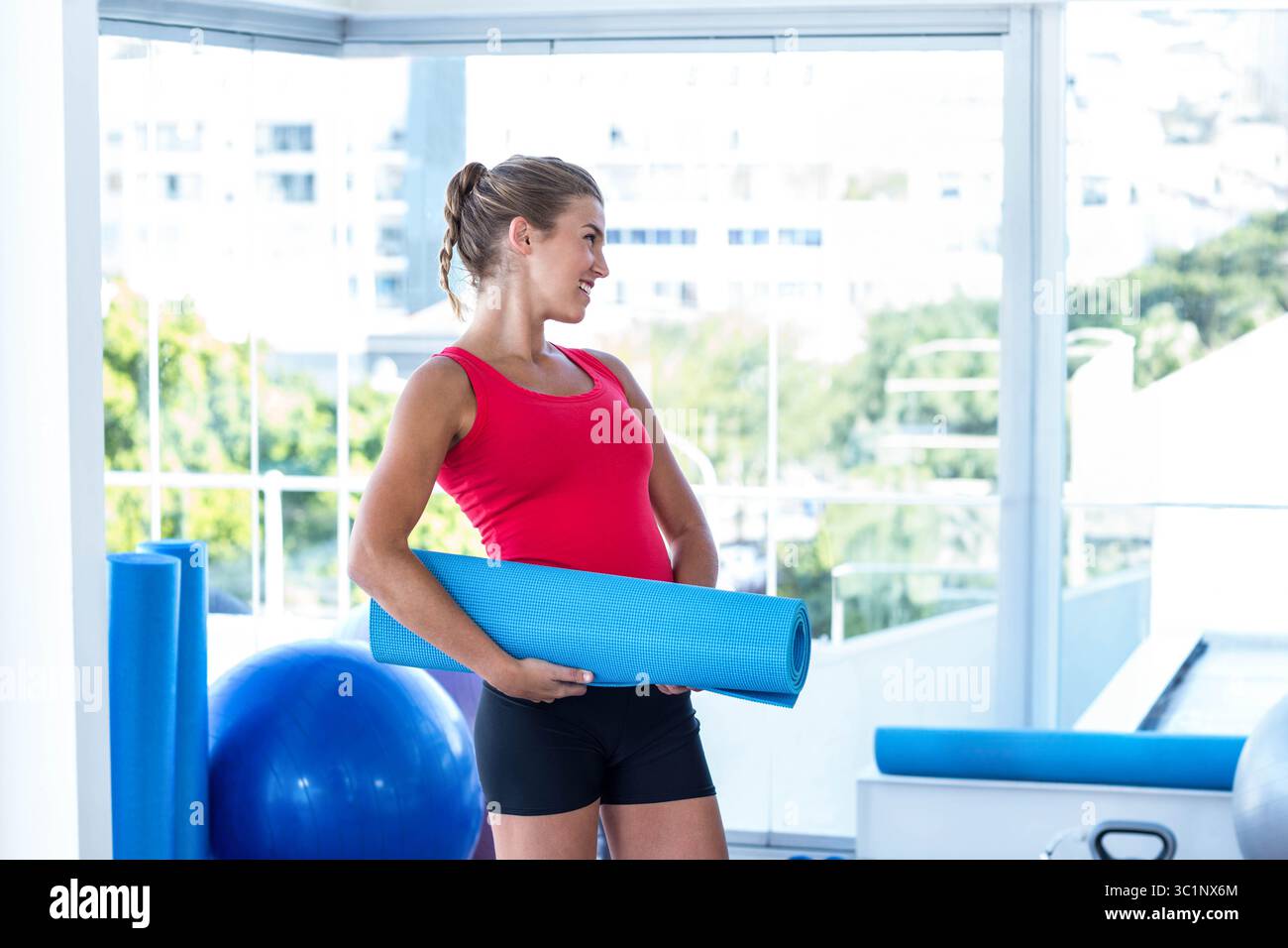 Woman holding rolled blue exercise mat in bright fitness studio with foam rollers and kettlebell Stock Photo