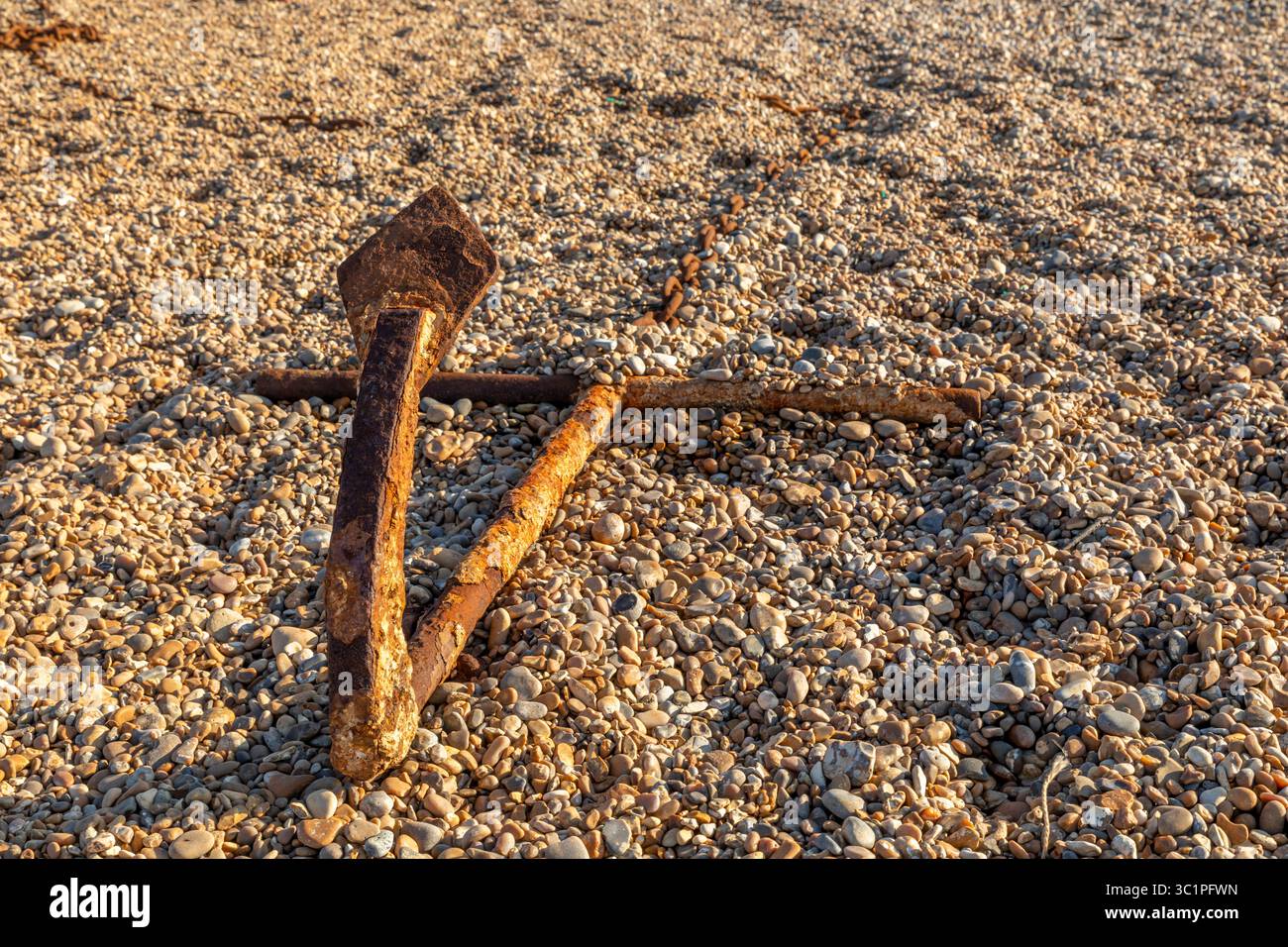 A rusty anchor lying on a shingle beach in warm evening light, weathered by time and tide along the British coast. Stock Photo