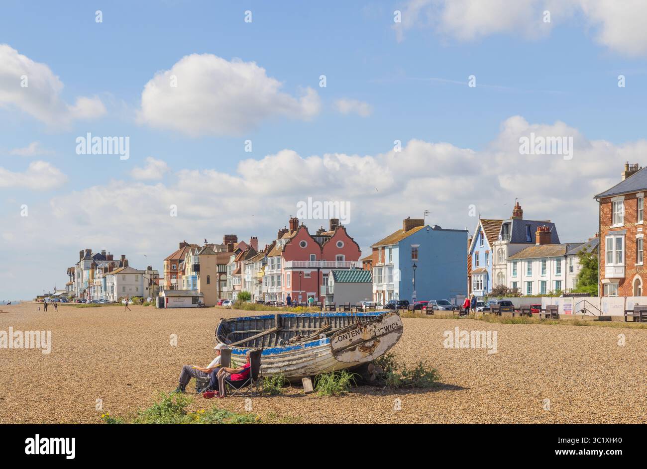 Aldeburgh, Suffolk. UK. View of the colourful buildings facing the beach, with tourists relaxing by an abandoned fishing boat in the foreground. Stock Photo