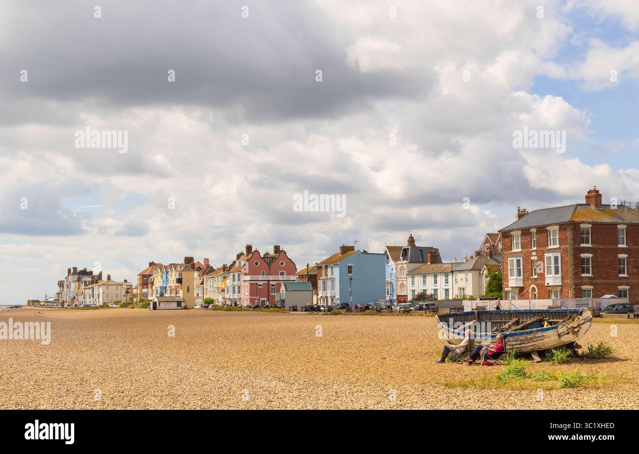 Aldeburgh, Suffolk. UK. View of the colourful buildings facing the beach, with tourists relaxing by an abandoned fishing boat in the foreground. Stock Photo