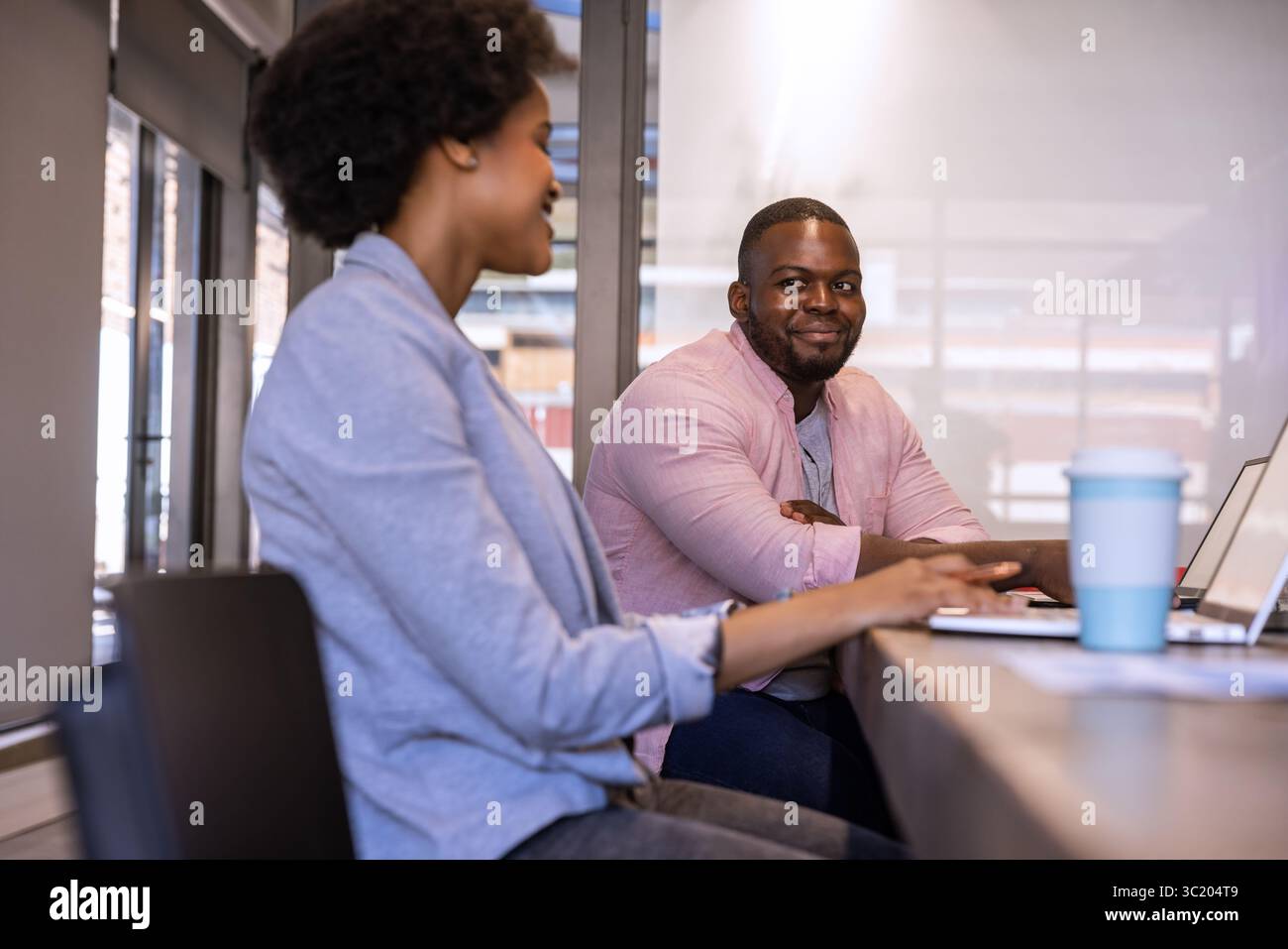 African American colleagues sitting in office discussing project using laptops with travel mug Stock Photo