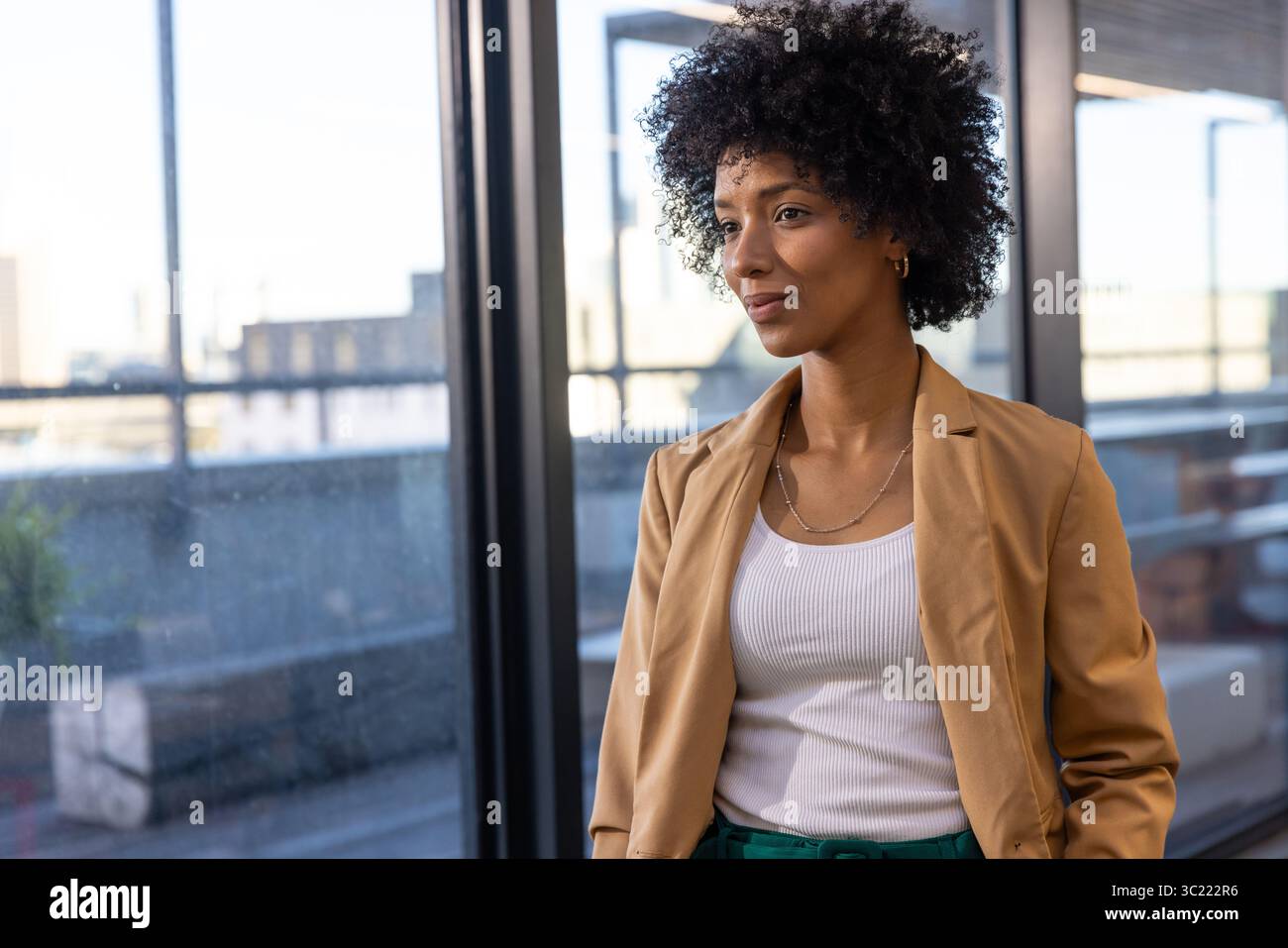 African American woman standing by window, metal railing in office wearing hoop earrings, necklace Stock Photo