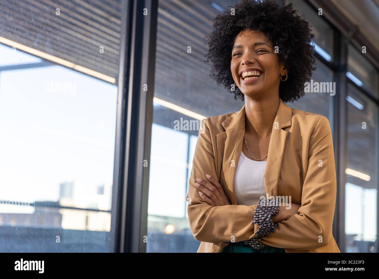 African American woman smiling by glass windows in office corridor wearing blazer, copy space Stock Photo