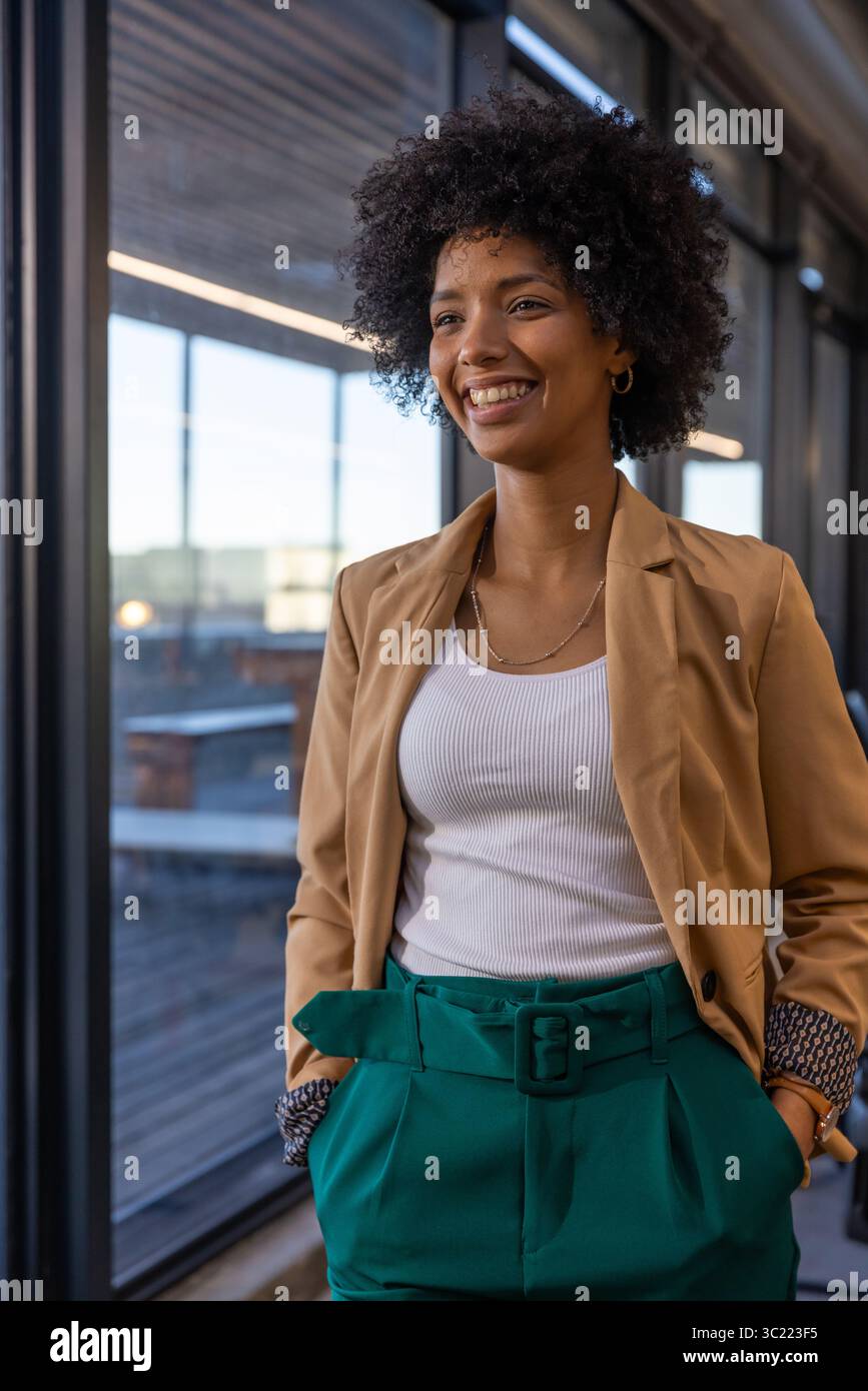 African American woman wearing hoop earrings and gold necklace standing in corridor by glass wall Stock Photo