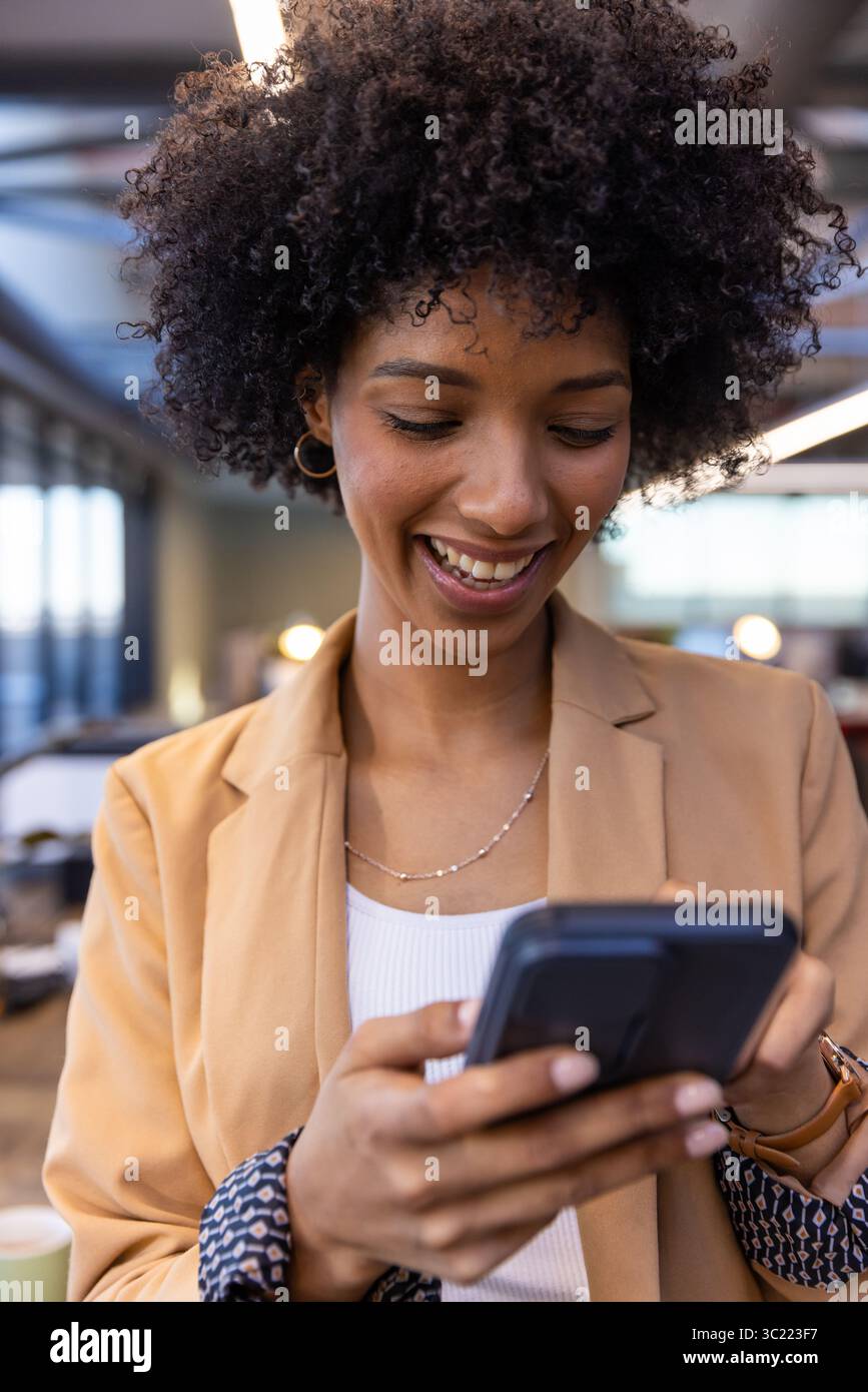 African American woman wearing tan blazer smiling holding smartphone and checking screen at office Stock Photo