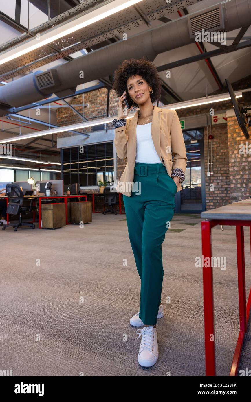 African American woman walking through open plan office talking on smartphone near red-framed desks Stock Photo