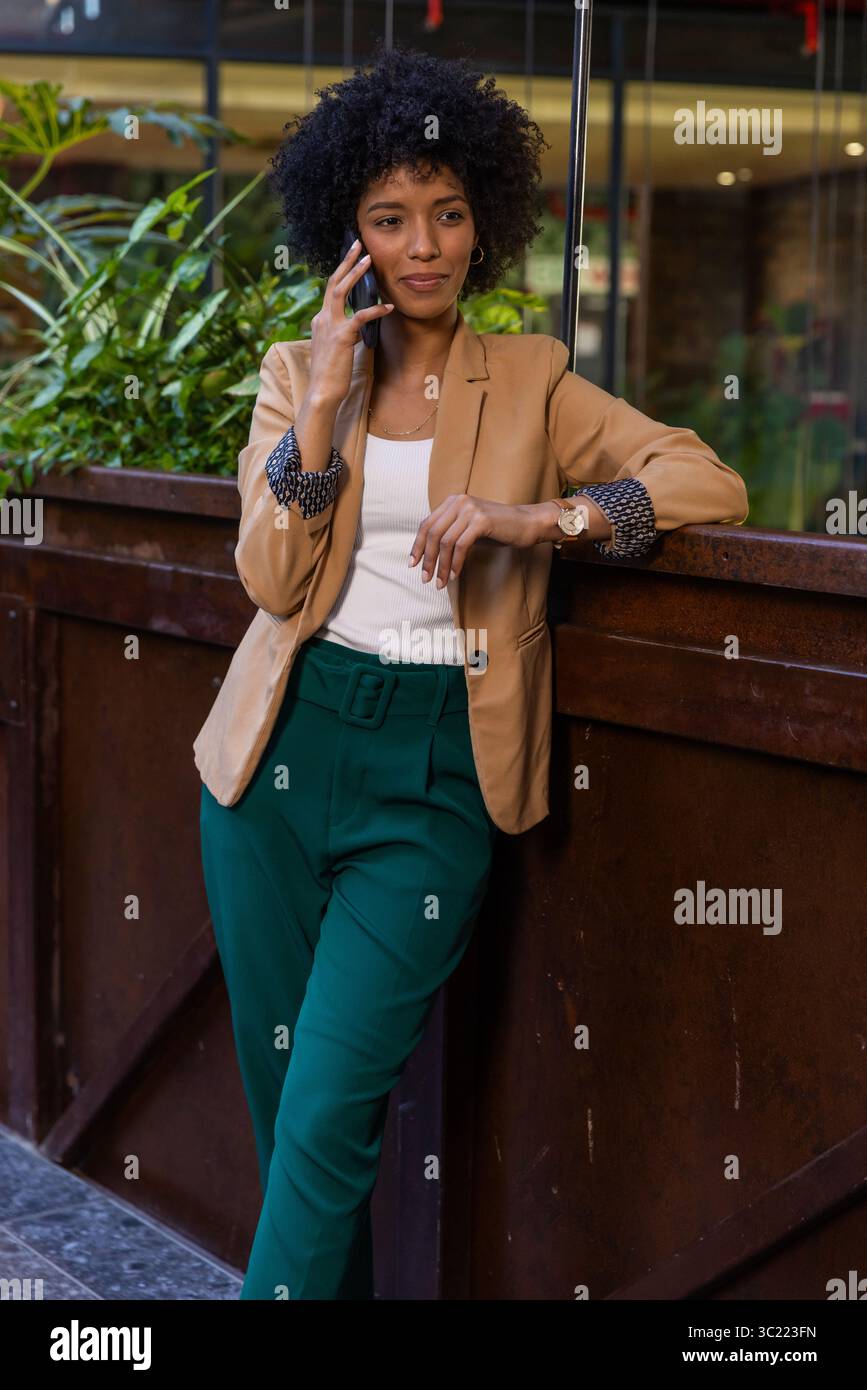 African American woman leaning on counter by wooden planter at office lobby talking on smartphone Stock Photo