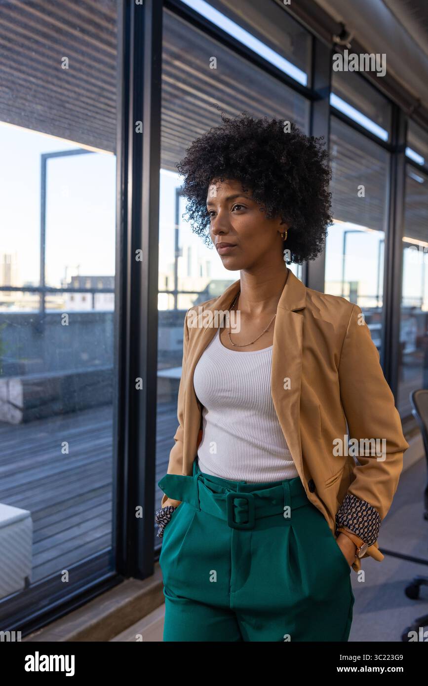 African American woman standing by windows in office wearing blazer, teal pants overlooking skyline Stock Photo