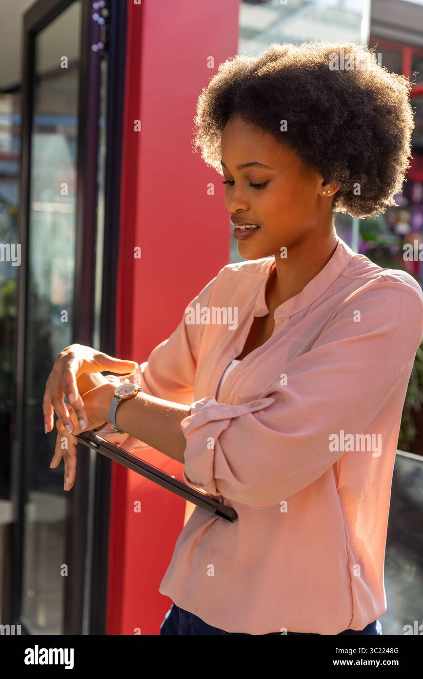 African American woman checking smartwatch and holding tablet at storefront in business attire Stock Photo