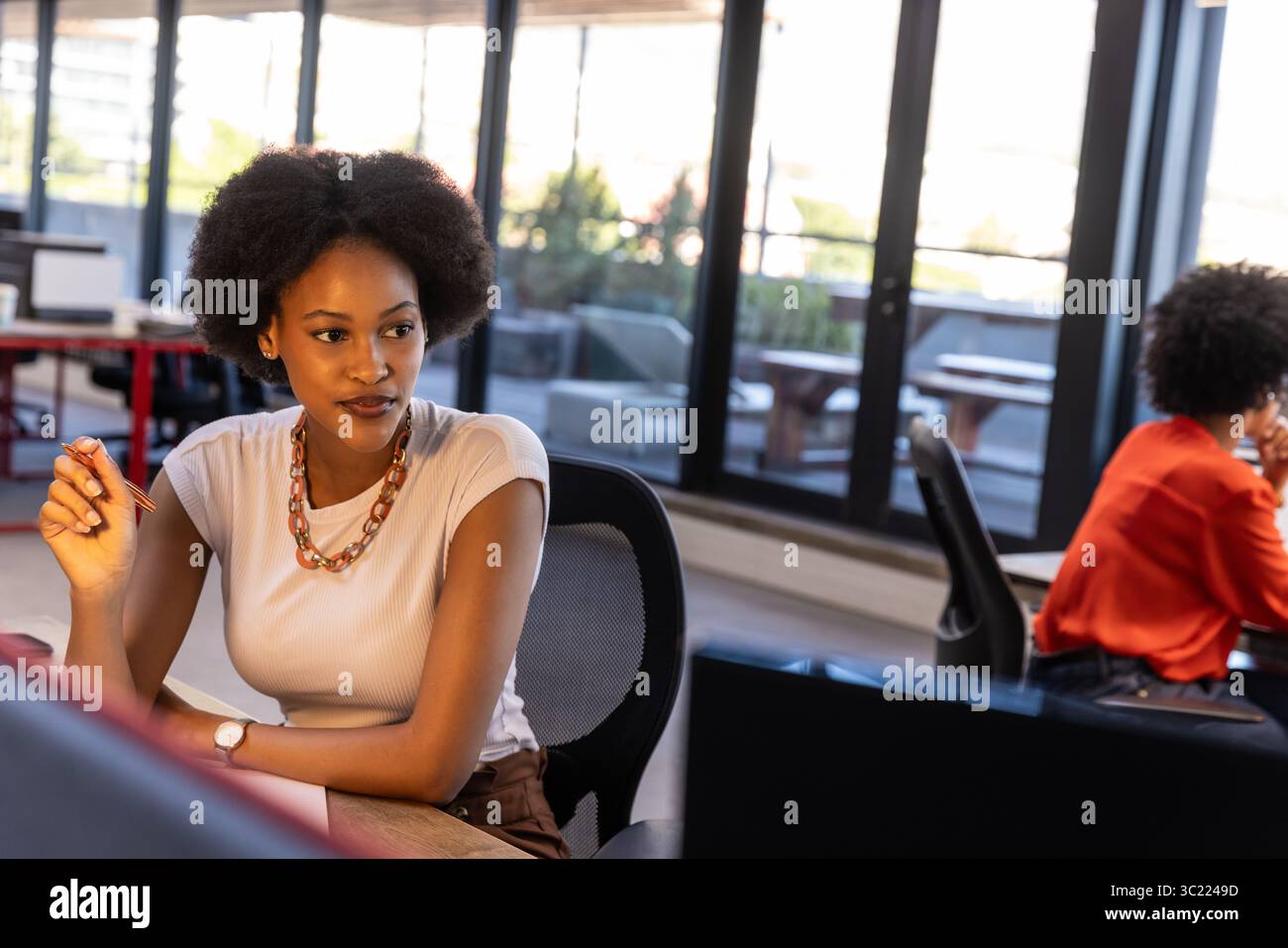 Diverse female coworkers sitting on ergonomic chairs at desk in office holding red pen by monitors Stock Photo