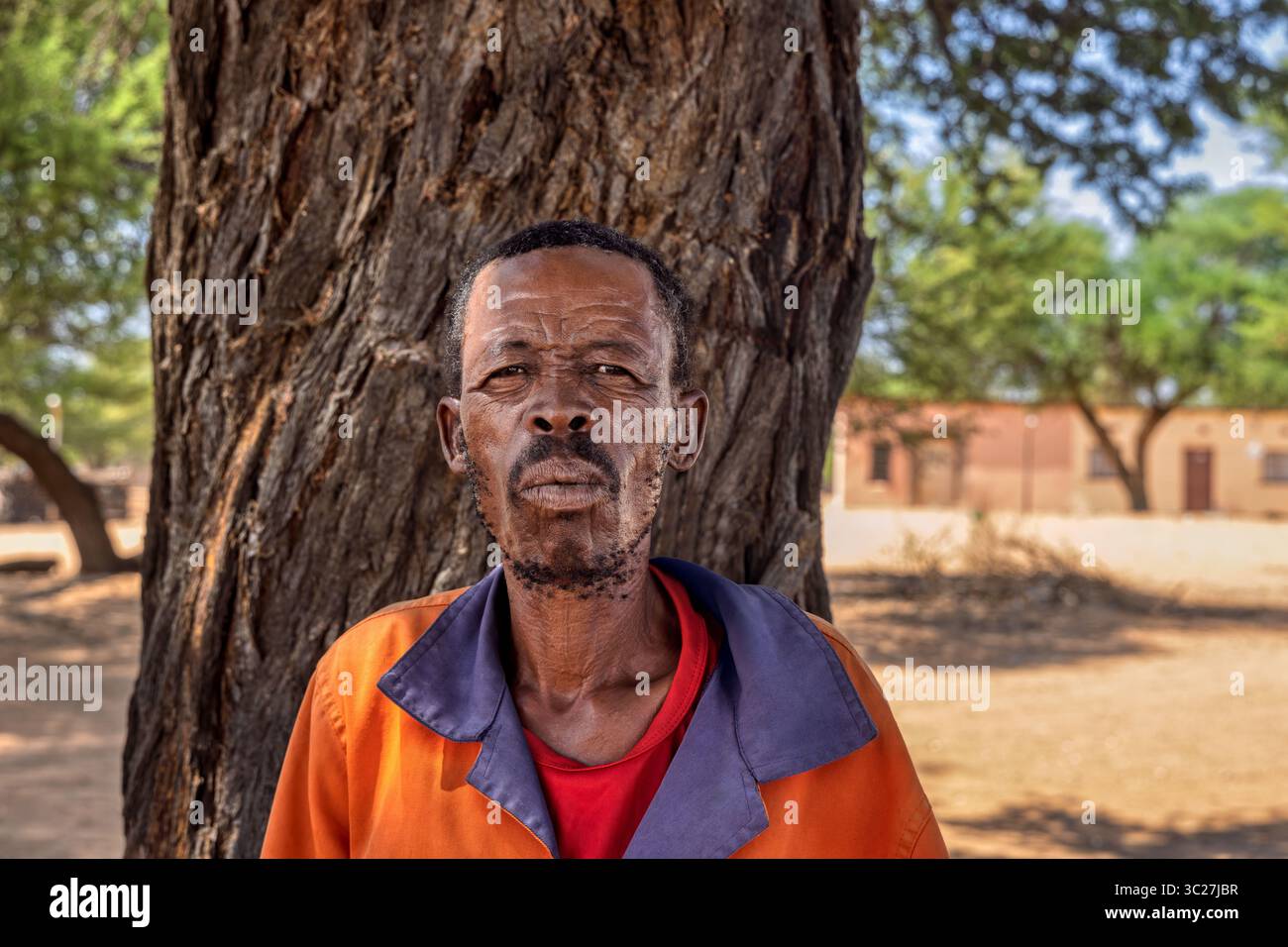 african village, old man wearing workwear , houses in the background rural, hard life standing under the shade of a tree Stock Photo