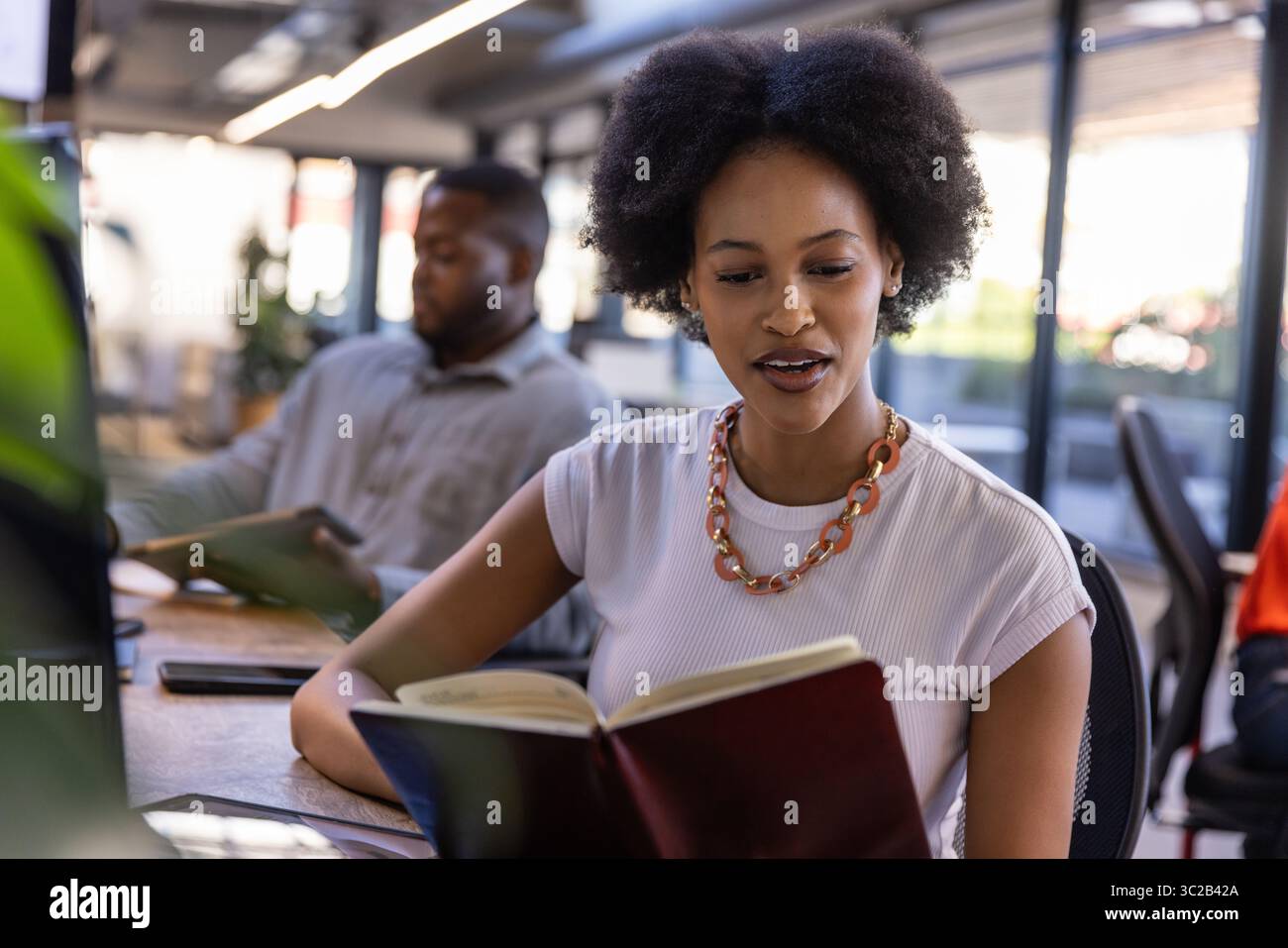 African American coworkers reading hardcover book and using tablet at office desks near windows Stock Photo