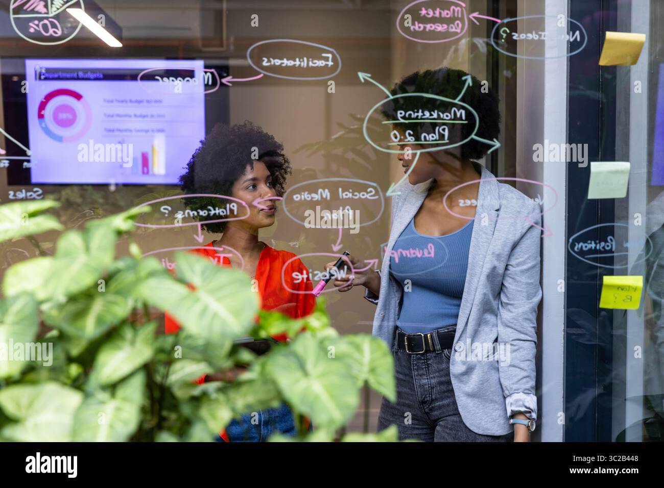 African American female coworkers sketching diagrams on glass wall at office with tablet and marker Stock Photo