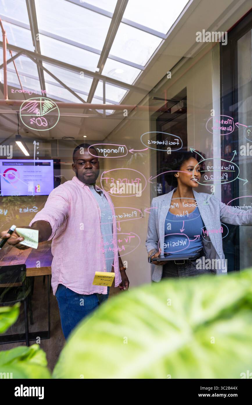 African American colleagues collaborating in office at glass partition with annotations using phone Stock Photo