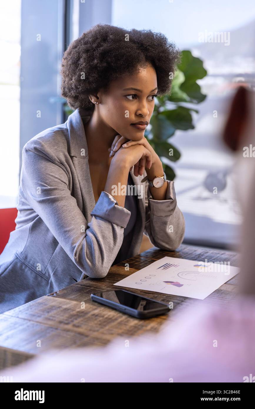 African American woman in grey blazer sitting at boardroom table reviewing chart with smartphone Stock Photo