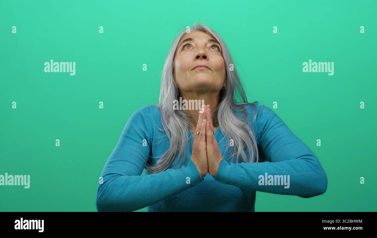 Senior woman with grey hair in prayer pose against a bright green ...