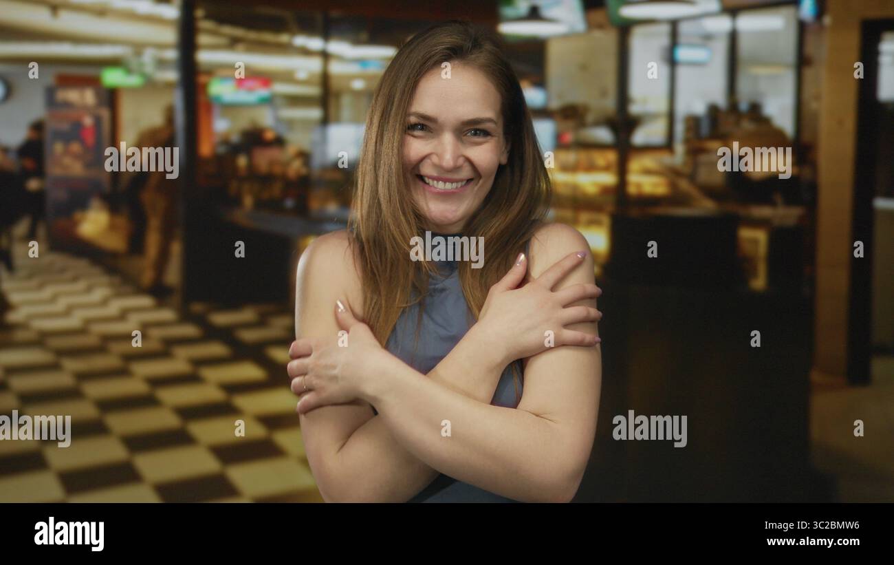 Smiling woman with long brown hair wraps arms around herself in building lobby with checkered floor under warm lights; inner peace. Stock Photo