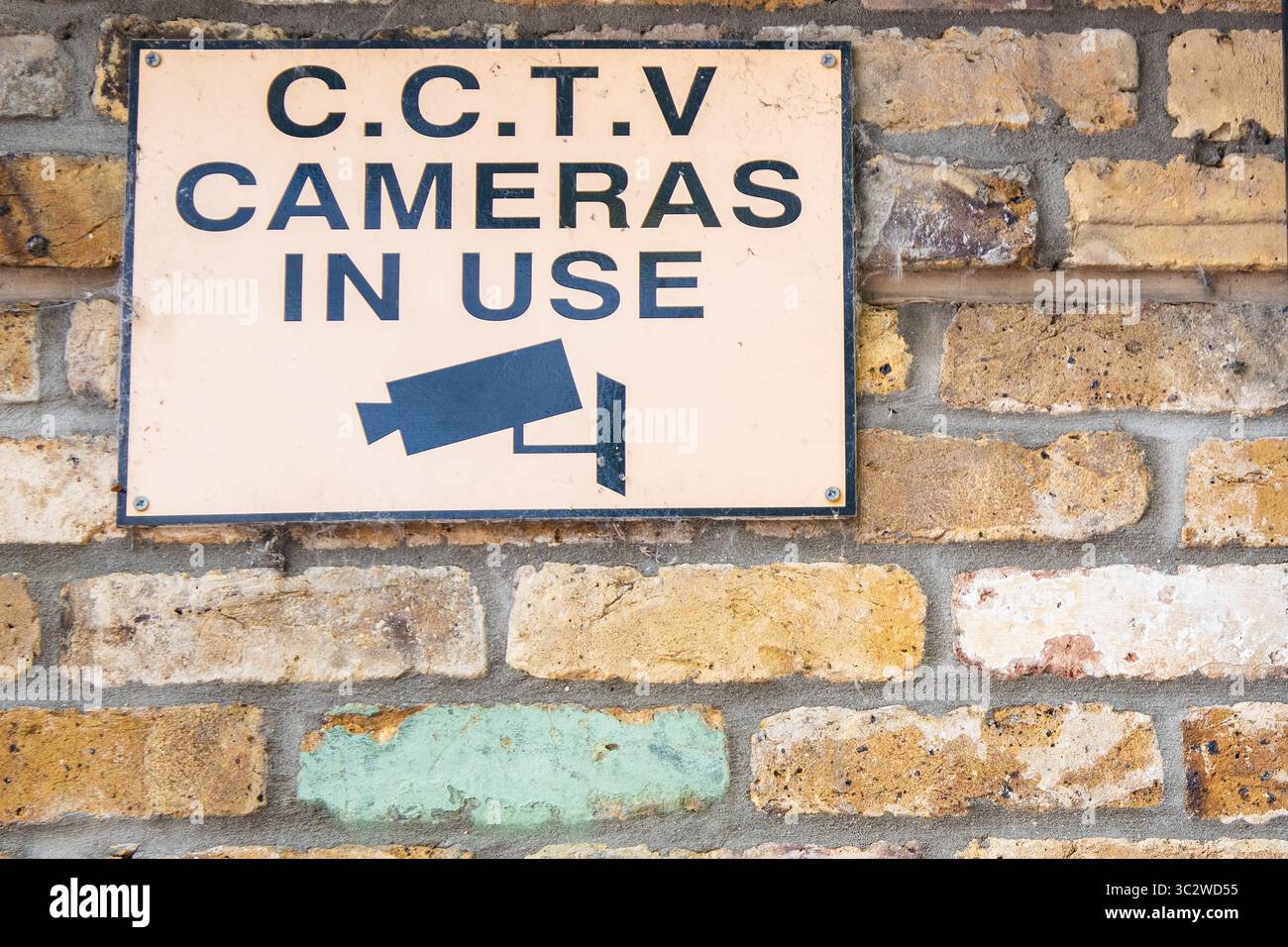 CCTV warning sign with black text on orange background and camera icon, mounted on the side of a brick building, indicating surveillance in operation. Stock Photo