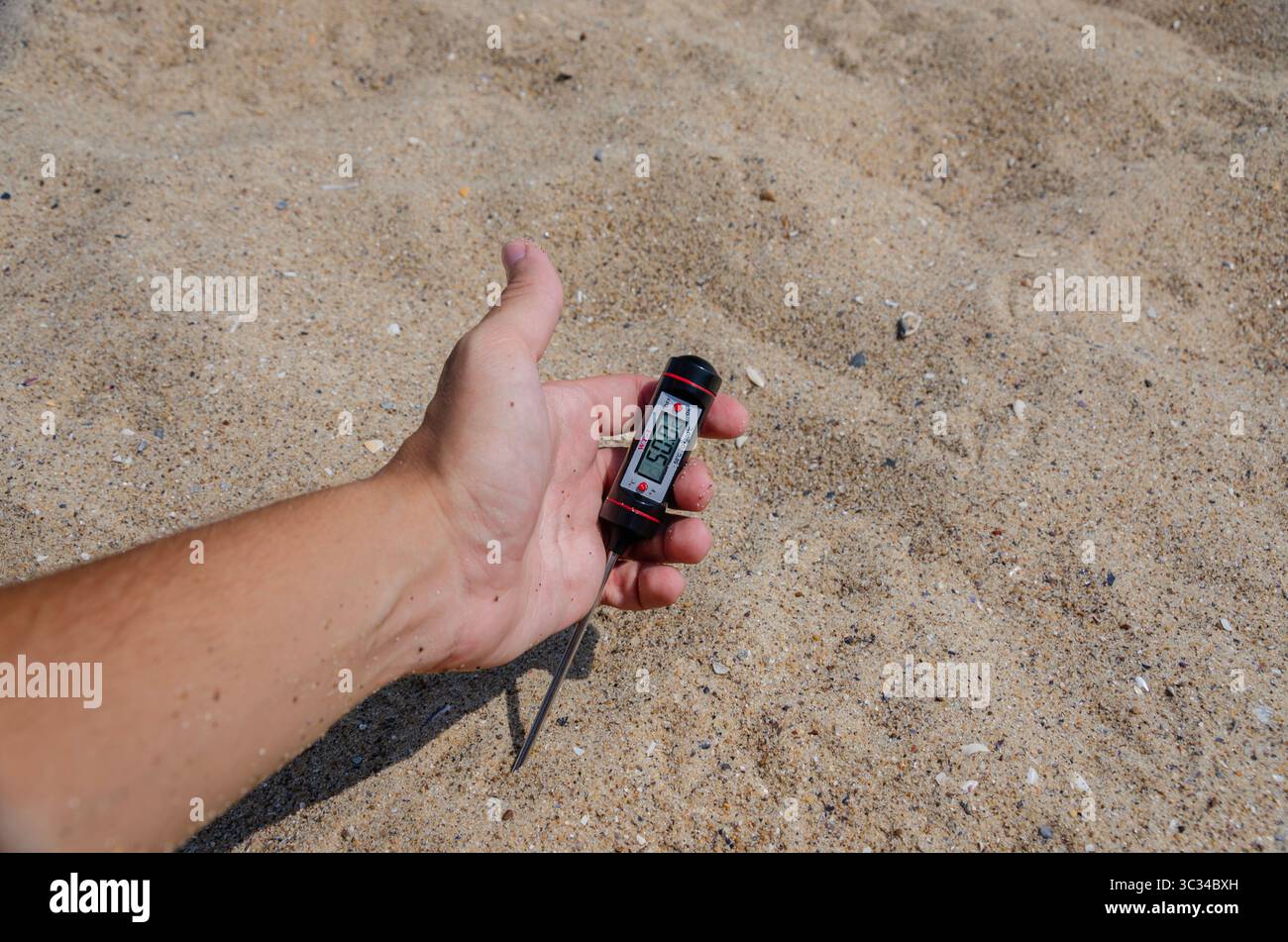 A close-up shot of a hand holding a digital thermometer, measuring the temperature of warm sand on a sunny beach, conveying concepts of summer heat, t Stock Photo