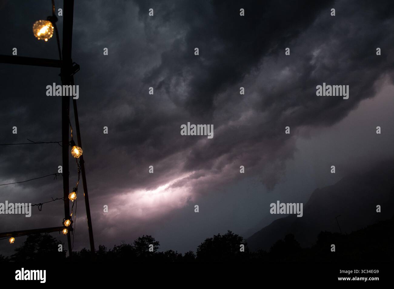 Dark storm clouds rolling over mountain peaks in Trentino, Italy, during a severe summer thunderstorm. Warm lights from outdoor string bulbs contrast Stock Photo