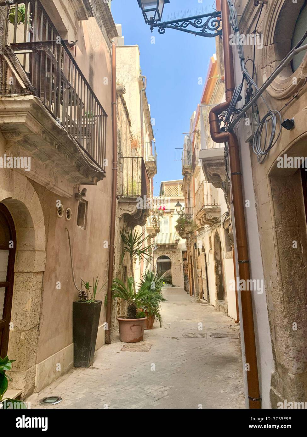 A charming slice of Ortigia, Syracuse — a narrow sunlit street framed by stone houses and quaint verandas, basking under a brilliant blue sky. Stock Photo