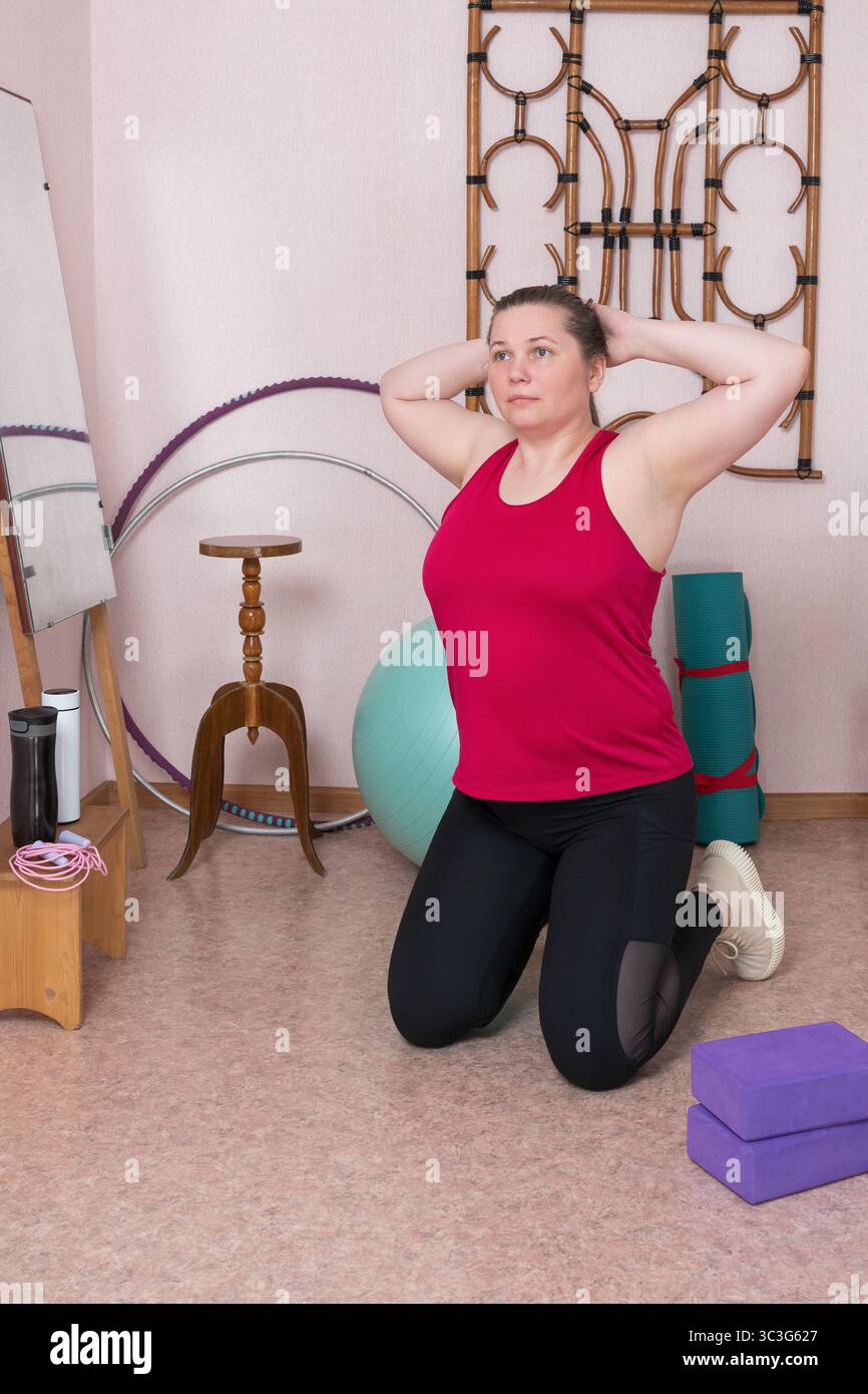 Middle aged woman is kneeling with her hands behind her head, doing an exercise during pilates workout at home. 44 year old woman is dressed in sports Stock Photo
