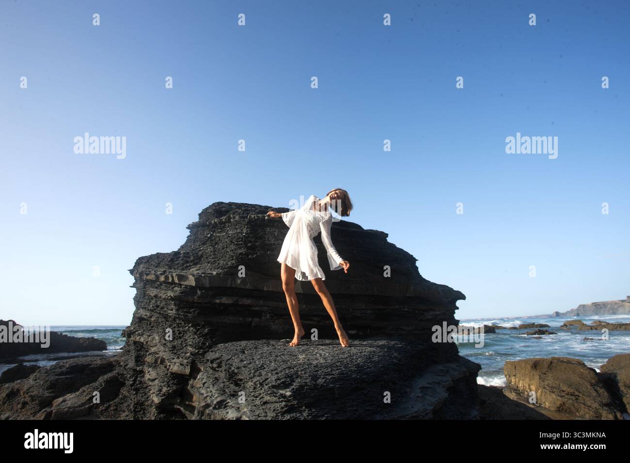 Beautiful natural woman in romantic simple white dress enjoying her beauty, while doing back band dancing pose in front of dark unique rock formation Stock Photo