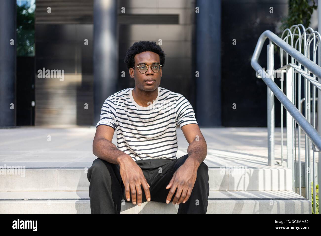 A young male student wearing glasses and a striped shirt sits on outdoor stairs, in front of a contemporary building, appearing focused and thoughtful Stock Photo
