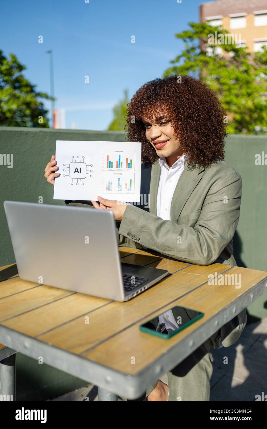 A trans woman in a suit sits at an outdoor table with a laptop, holding ...