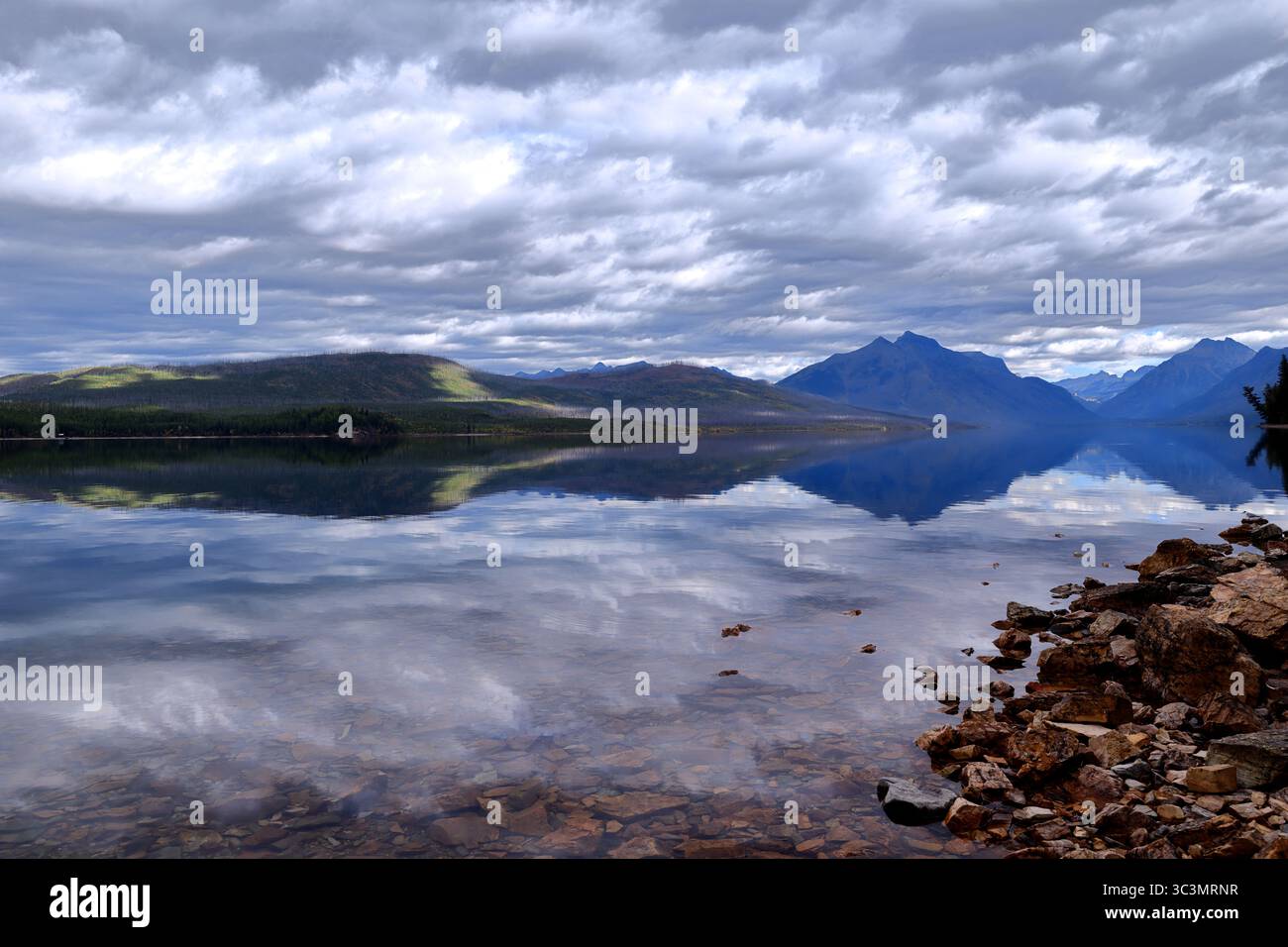 Rocky shore meets Lake McDonald's clear waters reflecting cloud-laden peaks and sunlit forested hills in Glacier National Park’s tranquil refuge. Stock Photo