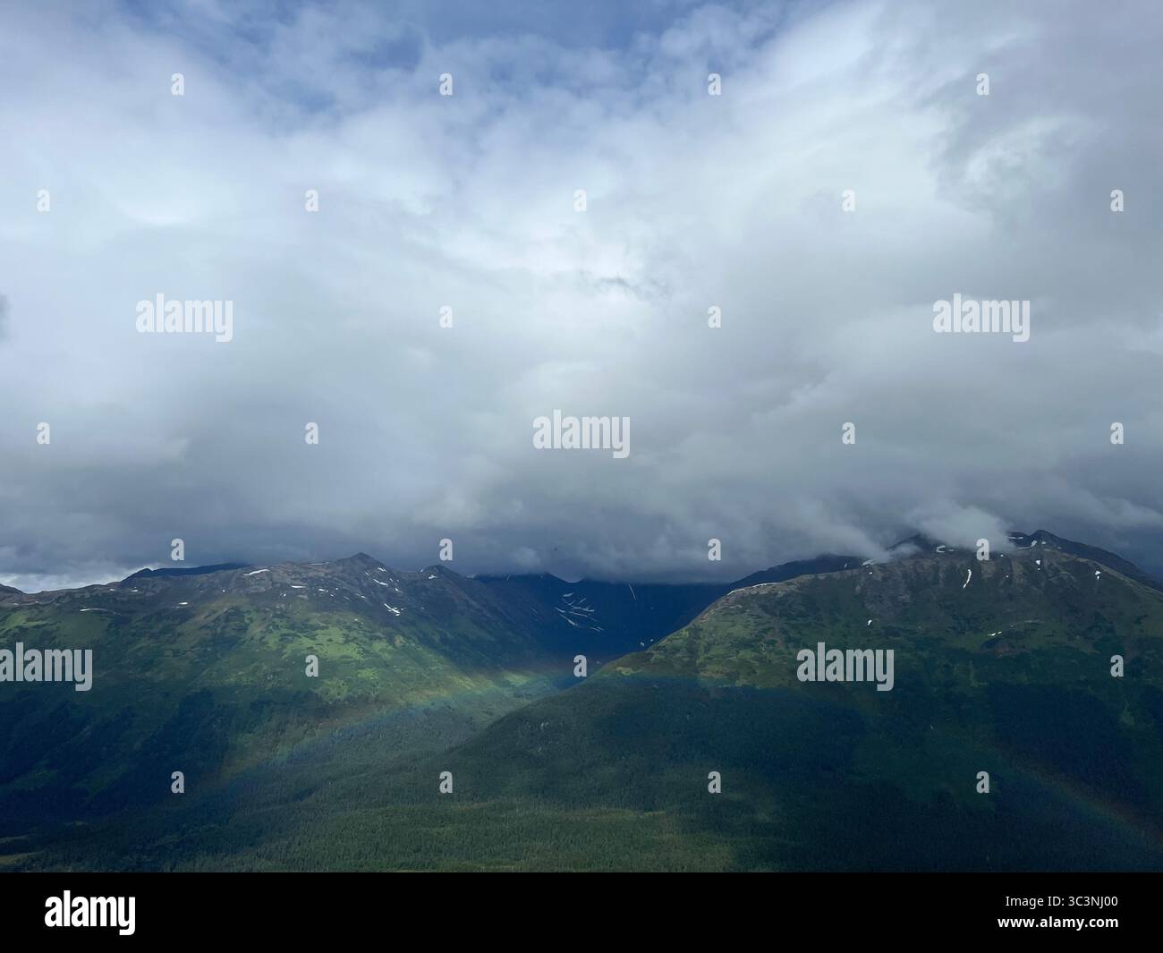Majestic Chugach Mountains, Alaska, under dramatic storm clouds with green slopes, snow, and a subtle rainbow. Rugged Alaskan wilderness - Smartphone Captured Stock Image