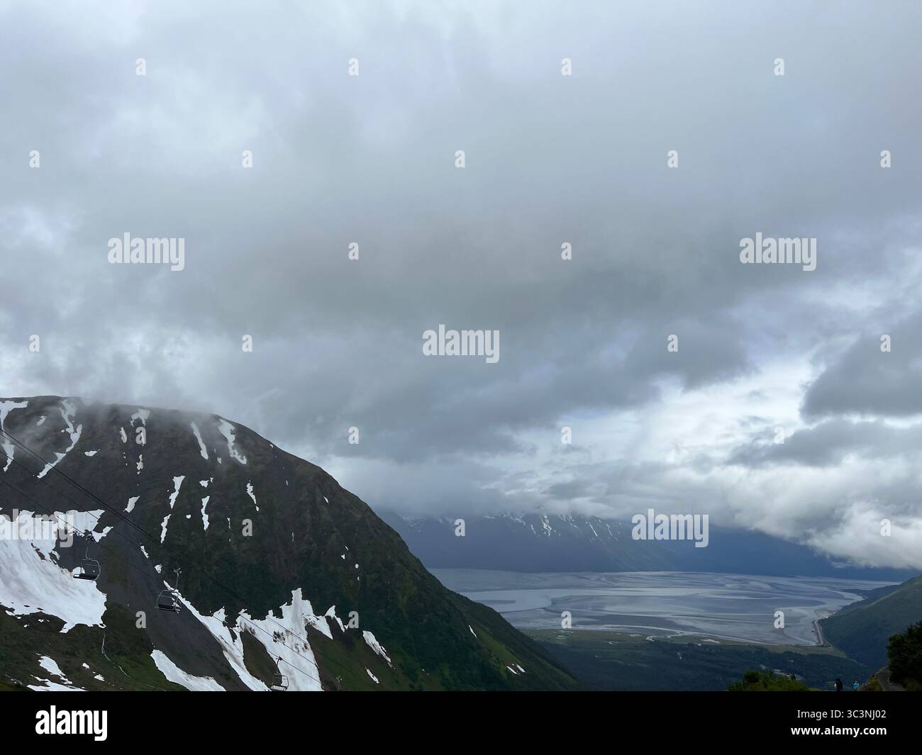 Dramatic snow-capped Chugach Mountains, Alaska, under heavy storm clouds, overlooking a vast valley and distant water. Untouched wilderness vista. - Smartphone Captured Stock Image
