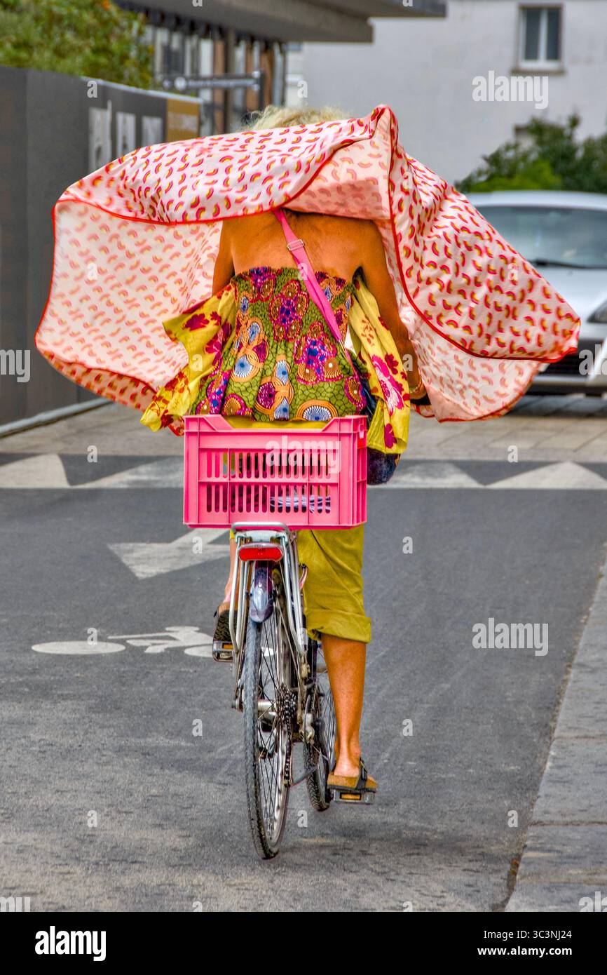 Woman in colourful clothes riding bicycle with rain cape blowing in wind - Tours, Indre-et-Loire (37), France. Stock Photo