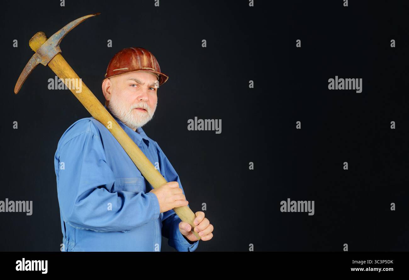 Serious builder in uniform with pickaxe on shoulder. Coal miner with pickax. Male laborer with hand tool for digging, mining. Construction worker in Stock Photo