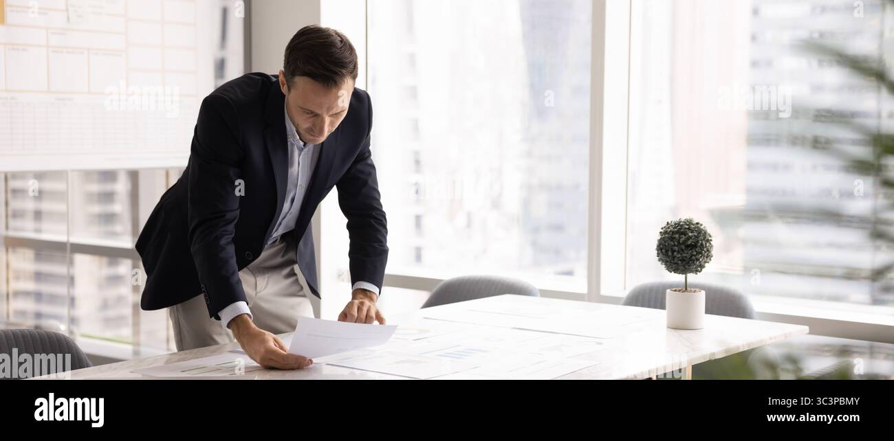 Businessman leaning over table, carefully examining papers with data Stock Photo