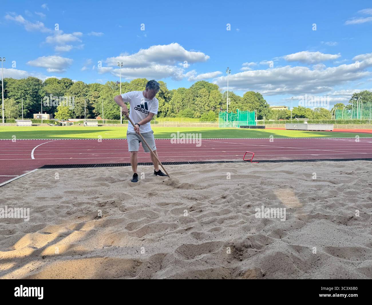 Man raking the sandpit at an athletics track during a sunny day in Lübeck, Germany. - Smartphone Captured Stock Image