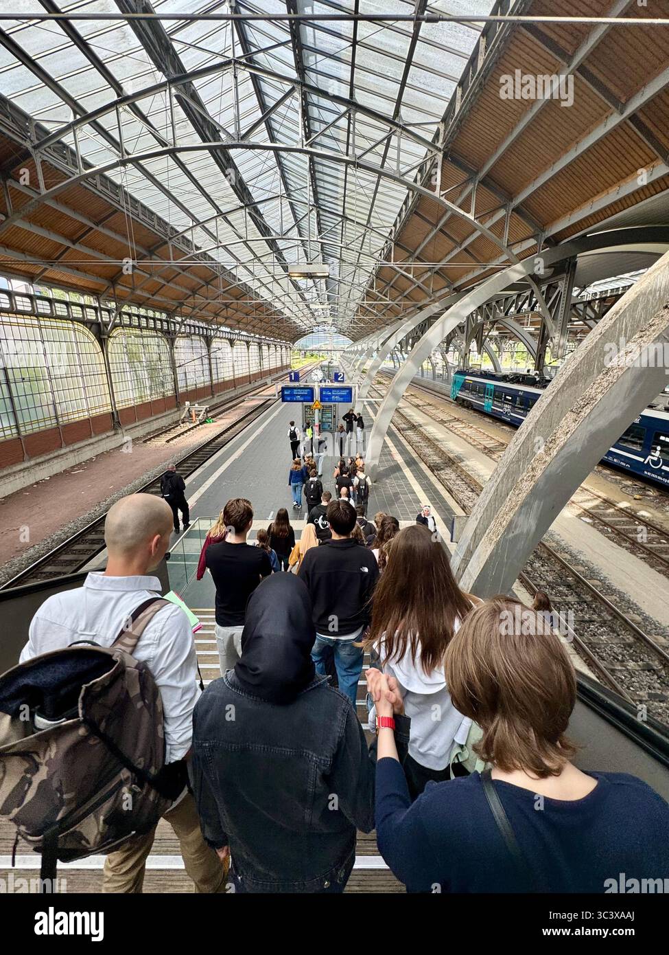 Passengers descending an escalator to the platform at Lübeck train station, Germany, under a large glass and steel canopy. - Smartphone Captured Stock Image