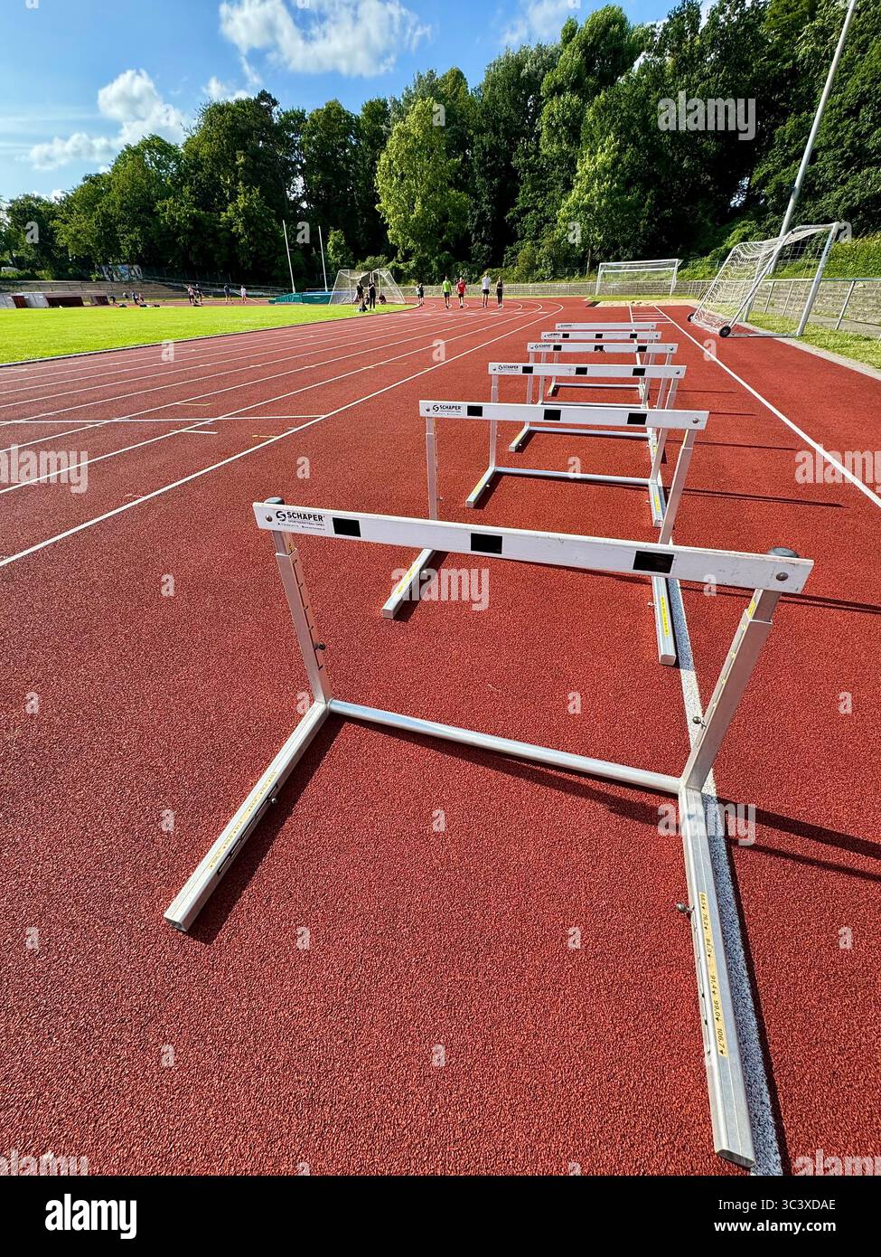 Hurdles lined up on a red athletics running track at a sports stadium in Lübeck, Germany, with athletes training in the background. - Smartphone Captured Stock Image